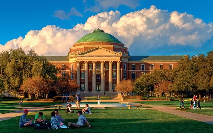 A photograph of the quad at Southern Methodist University, a large grassy area in front of Dallas Hall, pictured in the background. Students sit on the quad in groups of 4 or 5 people, while others cross it on foot.